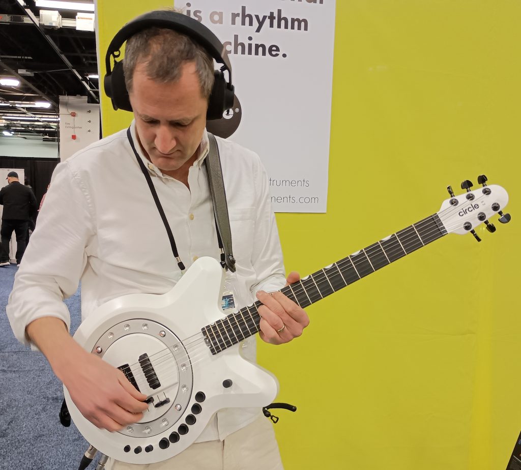 Man playing a white Circle Guitar in front of their yellow booth backdrop.