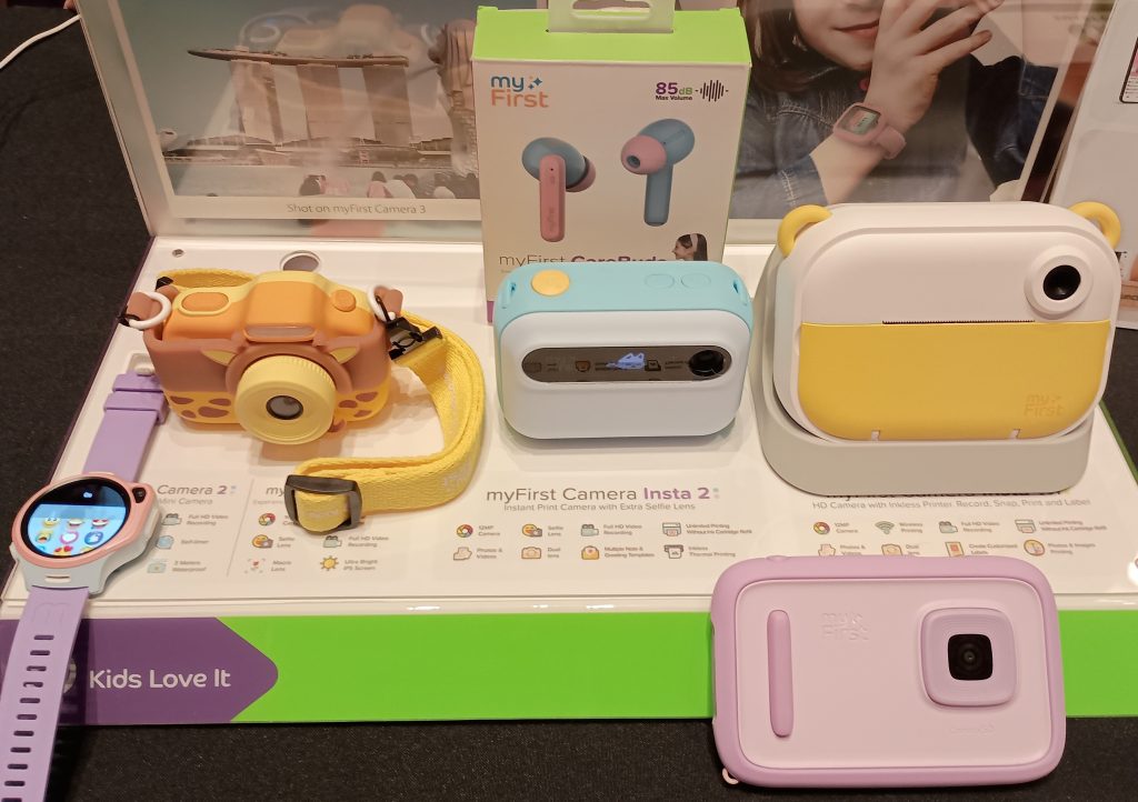 The myFirst product line of smart devices for kids. From left to right: purple banded watch, giraffe patterned camera, blue earbuds, blue instant camera, larger yellow instant camera, and pink digital camera.