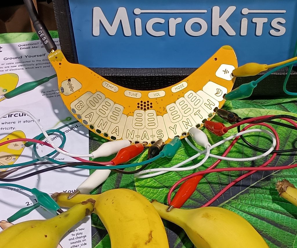 Yellow Banan-a-Synth modular synthesizer board shaped like a banana, with cords connecting it to real bananas, and MicroKits on a sign behind it.