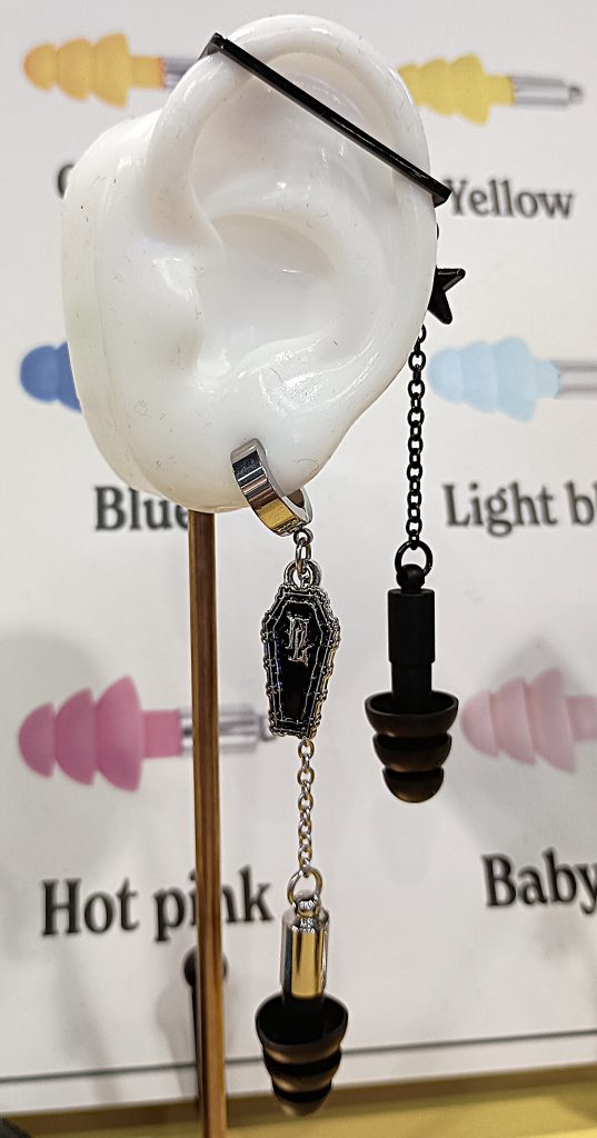 Black Plug & Play Jewelry earplugs dangling from 2 earrings on a white artificial ear, and a sign with different earplug colors in the background.